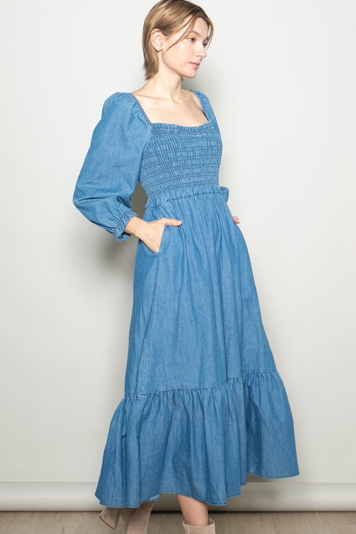 woman wearing a denim blue long sleeve midi dress with a smocked bodice and ruffle hem, standing with hands in pockets