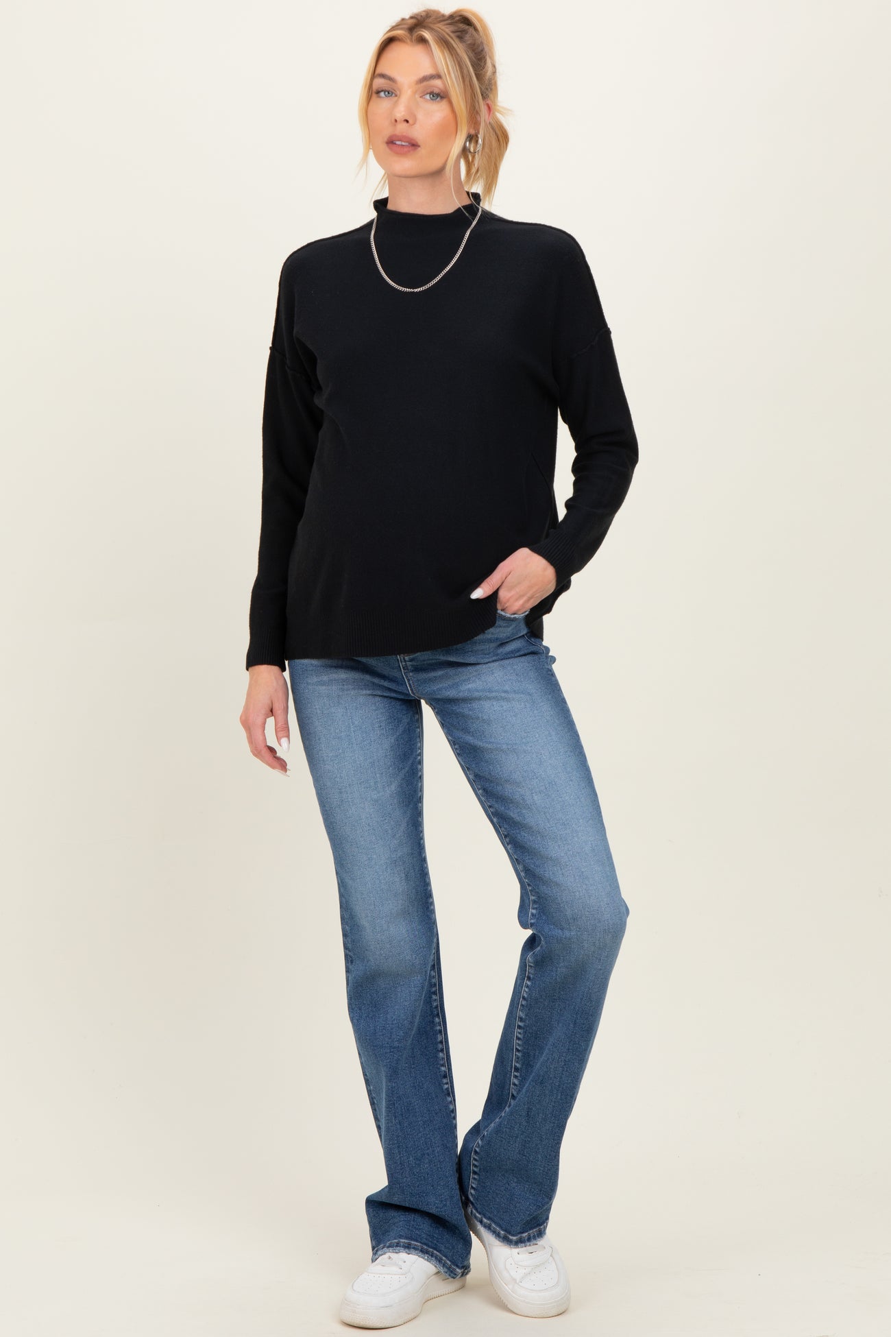 woman wearing a black maternity sweater and blue flared jeans, posing with one hand in pocket