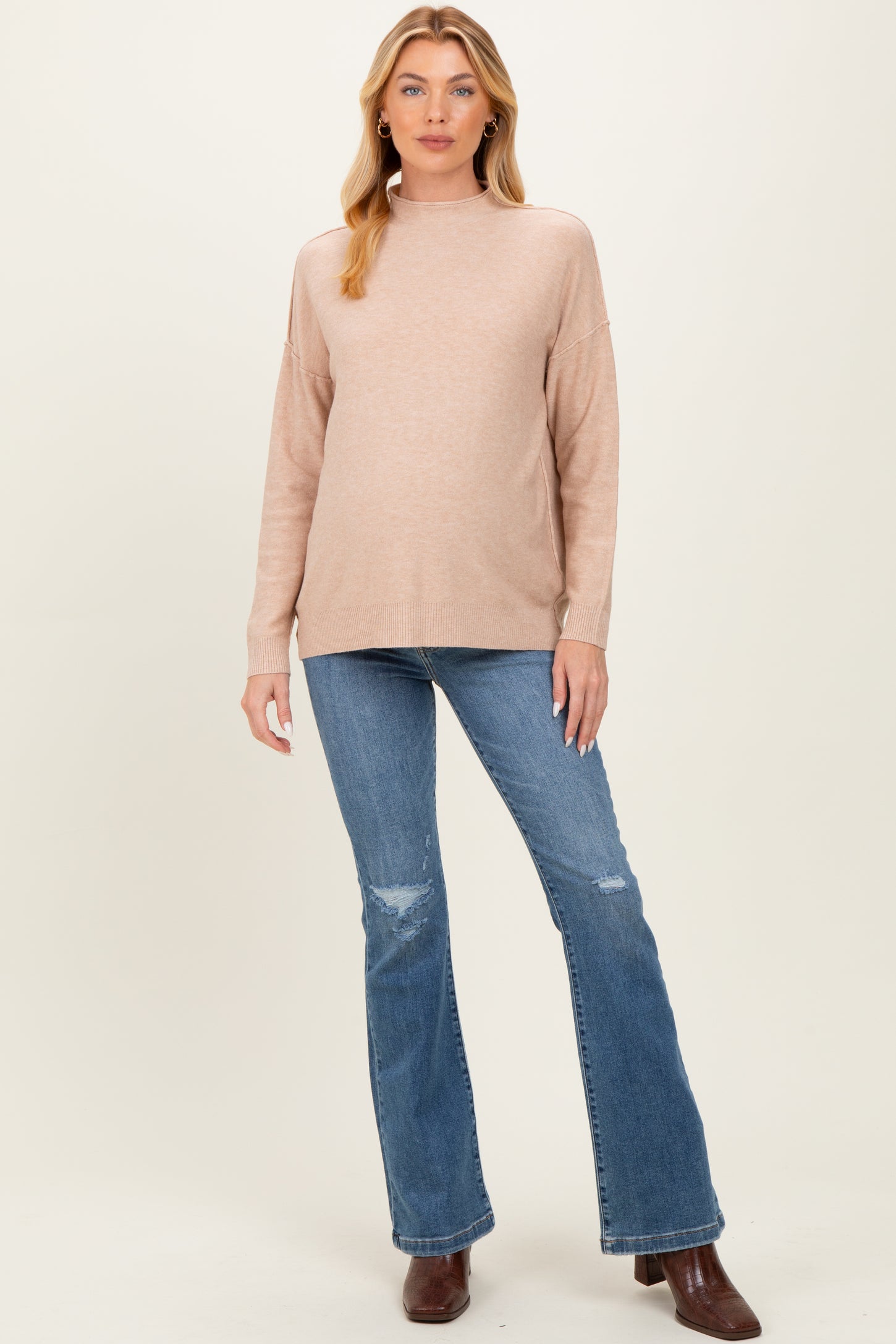 woman wearing a blush pink maternity sweater with a high neckline and distressed flared jeans, showing her baby bump