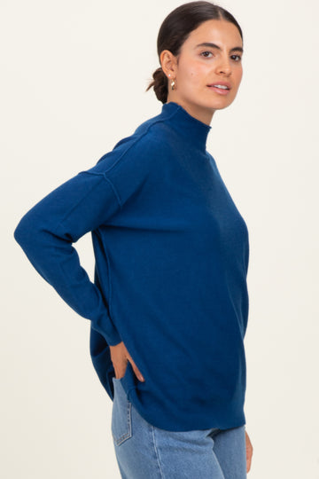 woman wearing a dark blue turtleneck sweater paired with light blue jeans, standing sideways against a neutral background