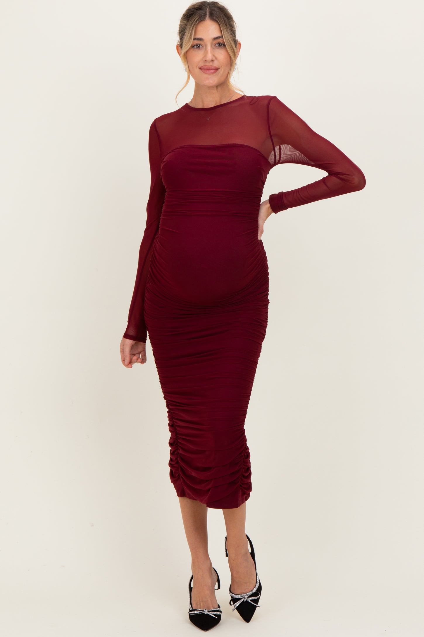 woman wearing a burgundy maternity dress with sheer long sleeves and ruched detailing, highlighting her baby bump