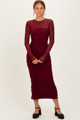 woman wearing a burgundy long sleeve midi dress with sheer neckline and ruched detailing