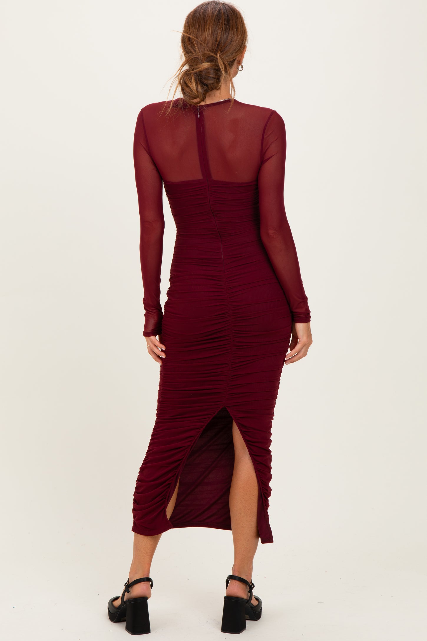 woman wearing a burgundy long sleeve ruched midi dress with a side slit and sheer long sleeves