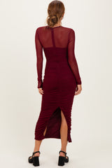 woman wearing a burgundy long sleeve ruched midi dress with a side slit and sheer long sleeves