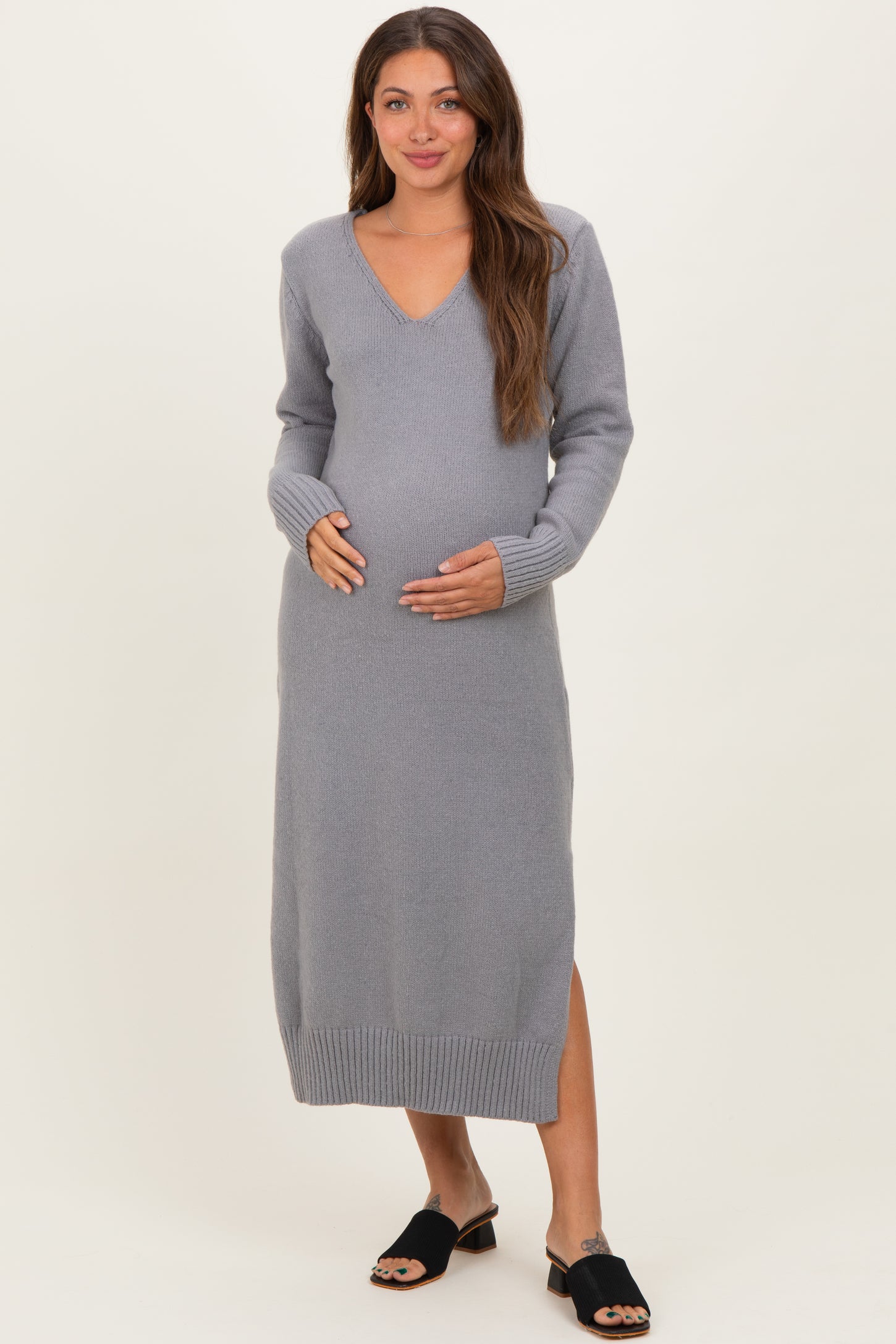 woman wearing a gray long sleeve maternity sweater dress with a v-neck and side slit, cradling her baby bump