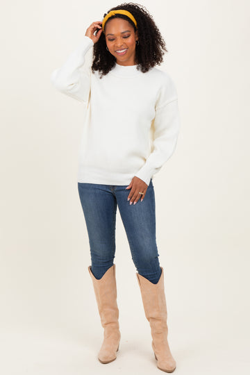 woman wearing a white oversized sweater, blue jeans, and beige knee-high boots, smiling with a yellow headband in her curly hair