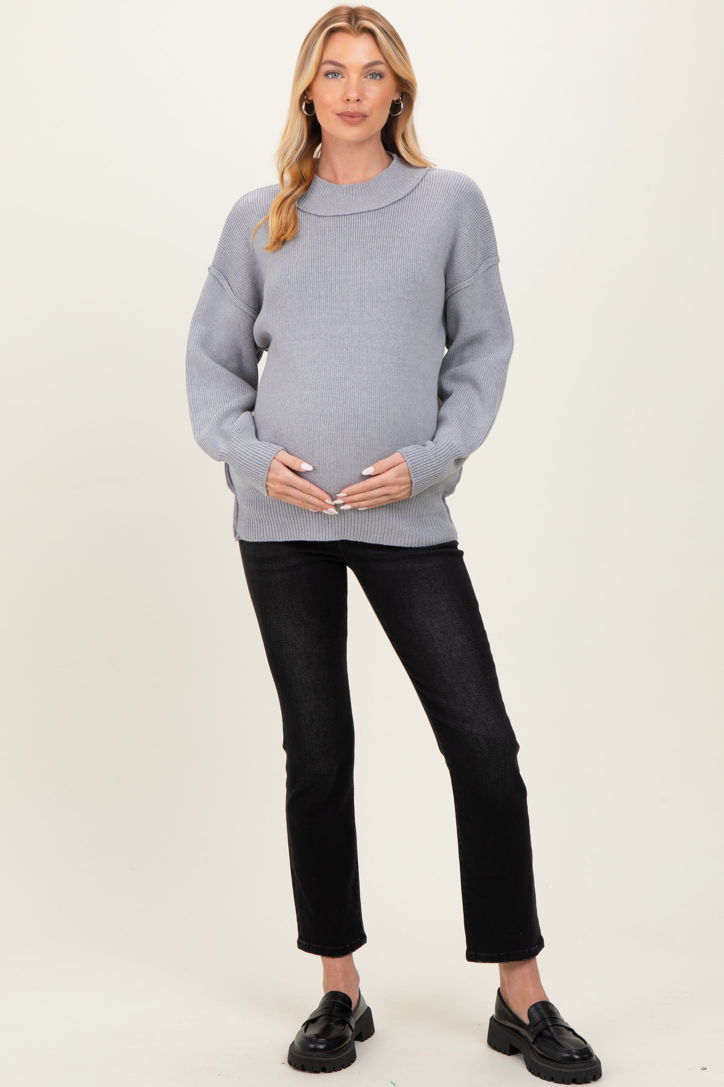 woman wearing a light gray maternity sweater and holding her baby bump with black jeans and loafers