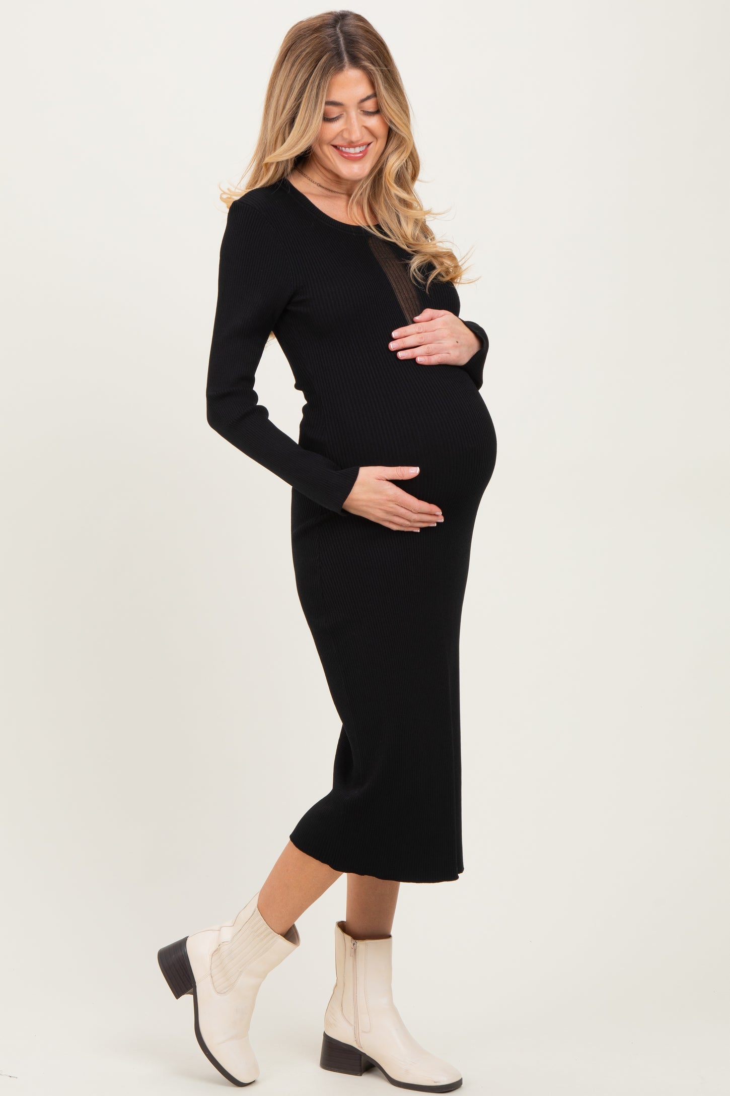 woman wearing a black ribbed maternity dress and cradling her baby bump, paired with white ankle boots
