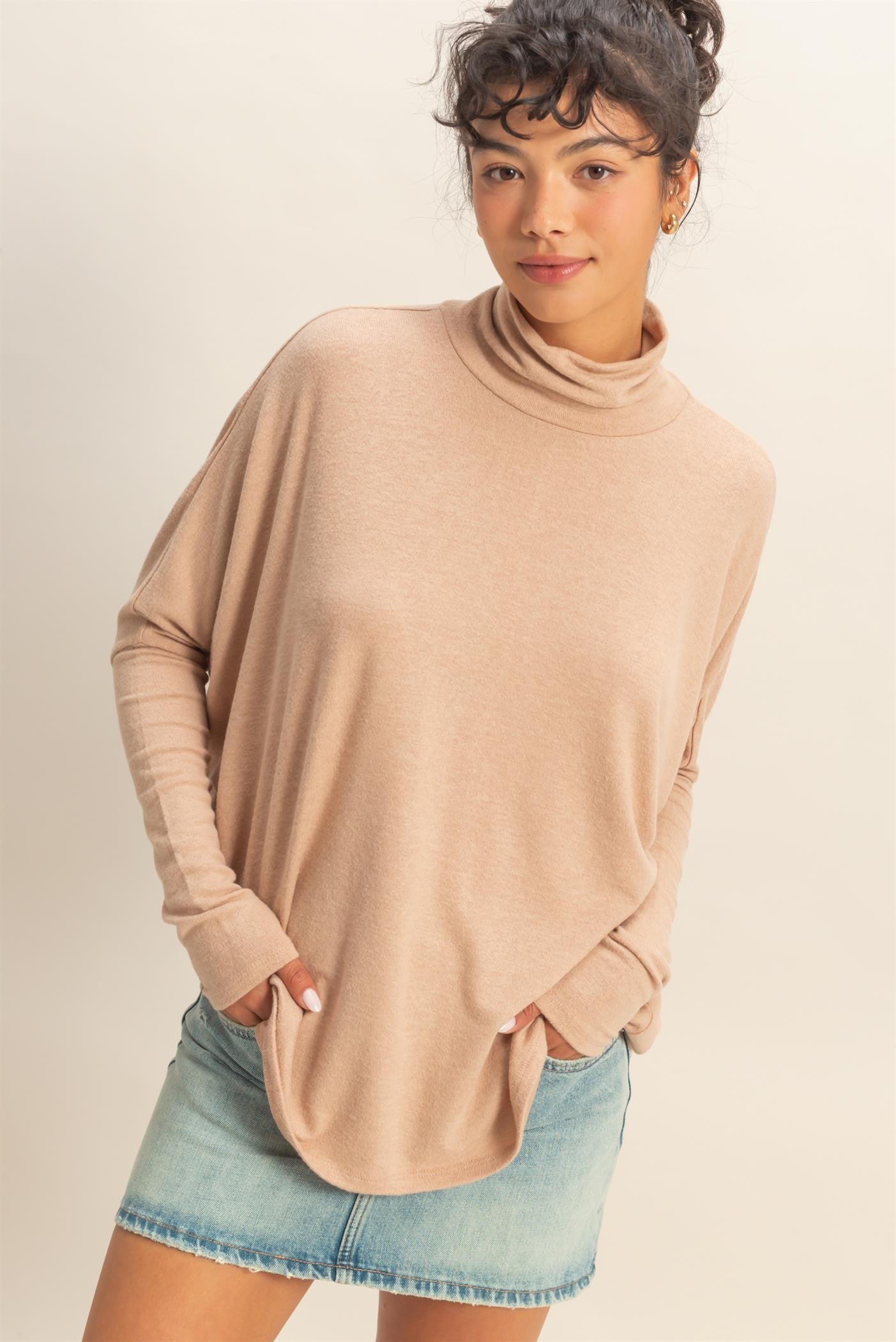 woman wearing a beige long sleeve turtleneck top with an oversized fit and front pockets, paired with a denim mini skirt