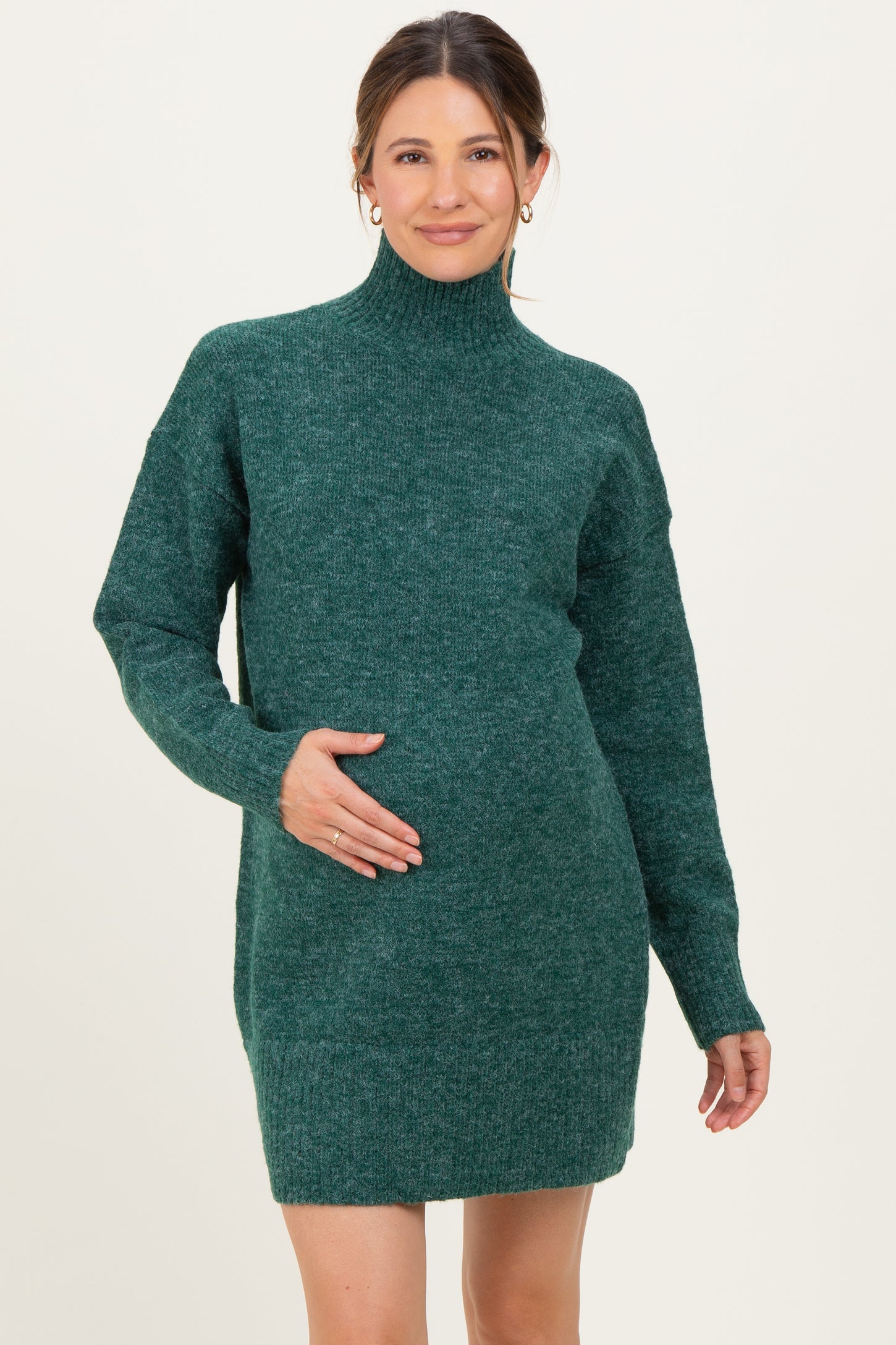 woman wearing a dark green maternity sweater dress with a high neck and relaxed fit, holding her baby bump