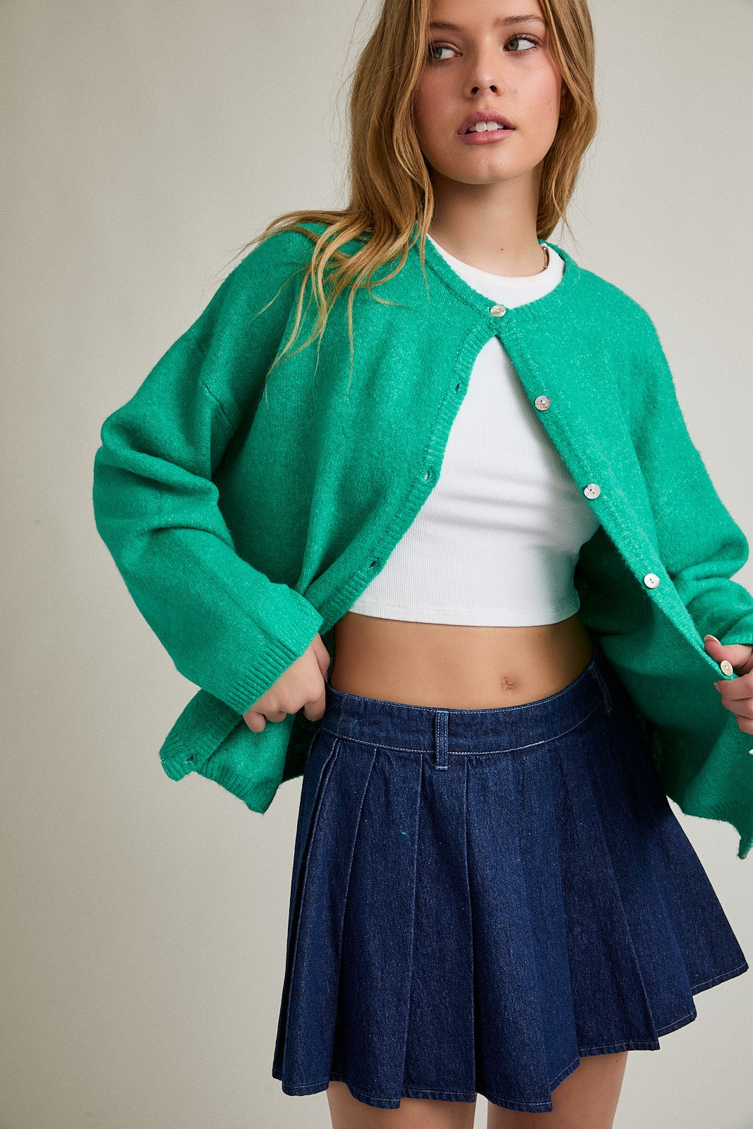 woman wearing a green cardigan over a white crop top and a denim pleated skirt