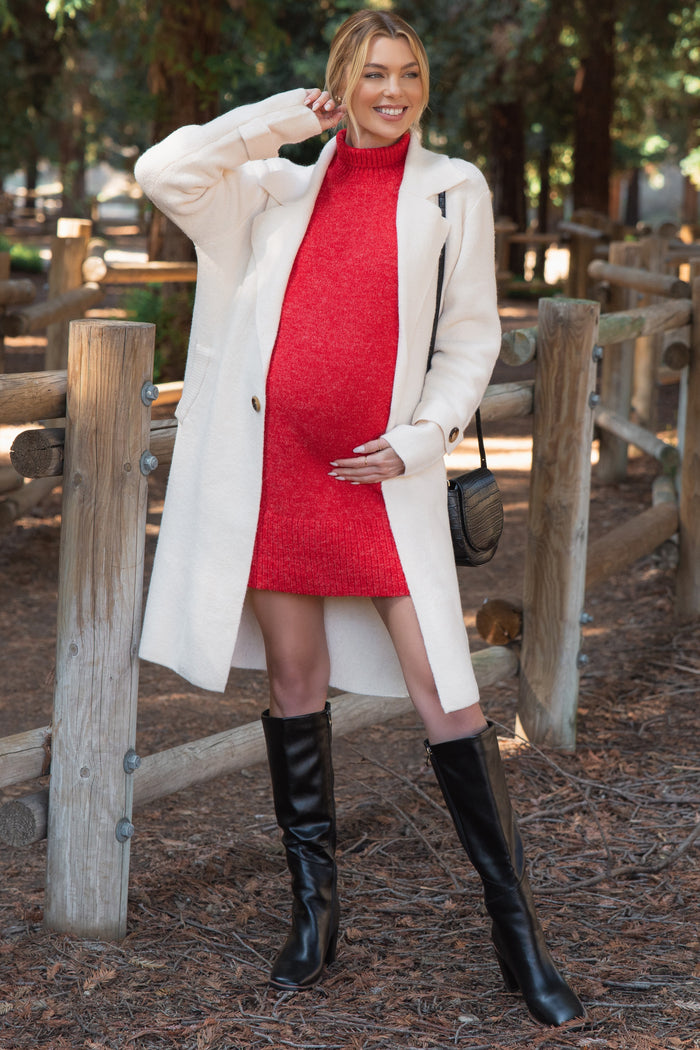 woman wearing a red knit maternity dress and a white coat, holding her baby bump in a forest setting