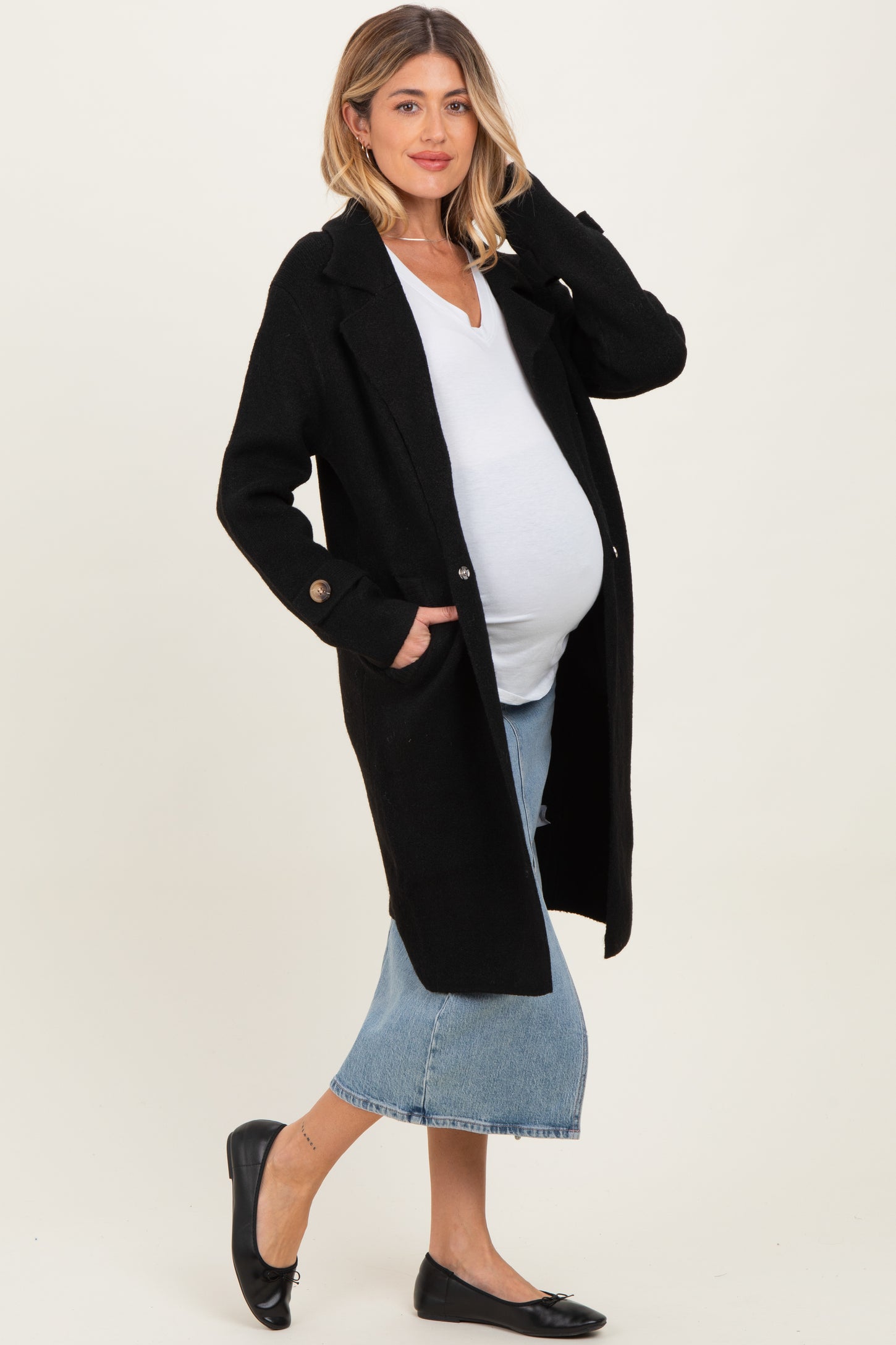 woman wearing a black maternity coat over a white v-neck top and blue denim skirt, showing her baby bump