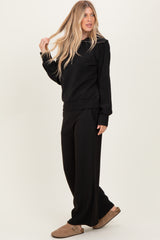 woman wearing a black ribbed sweater and matching wide-leg pants, styled with brown slippers and long blonde hair