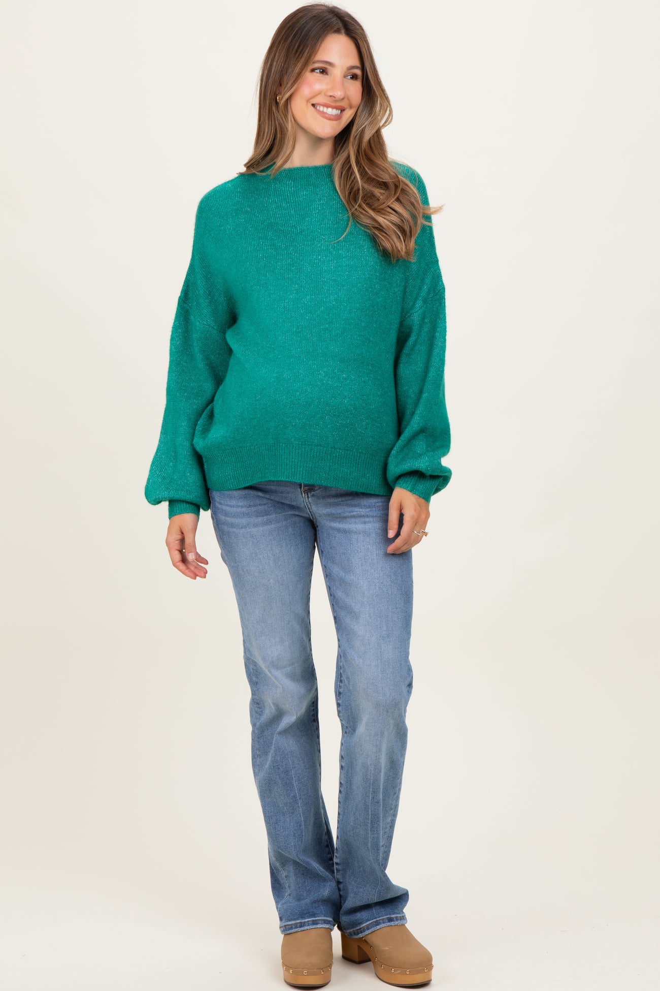 woman wearing a teal maternity sweater with balloon sleeves and light blue jeans, smiling at the camera