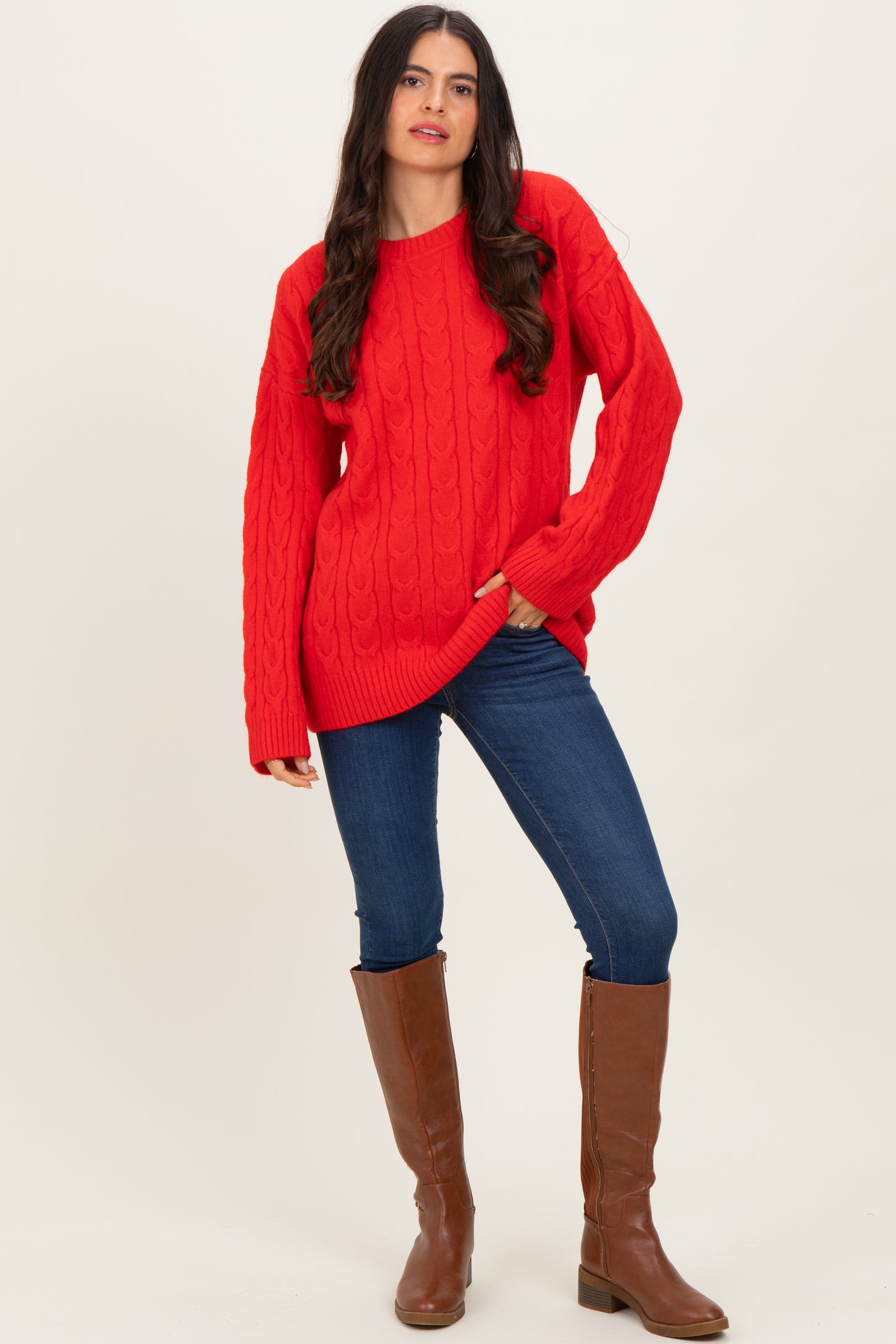 woman wearing a bright red cable knit sweater with long sleeves and blue skinny jeans, paired with brown knee-high boots