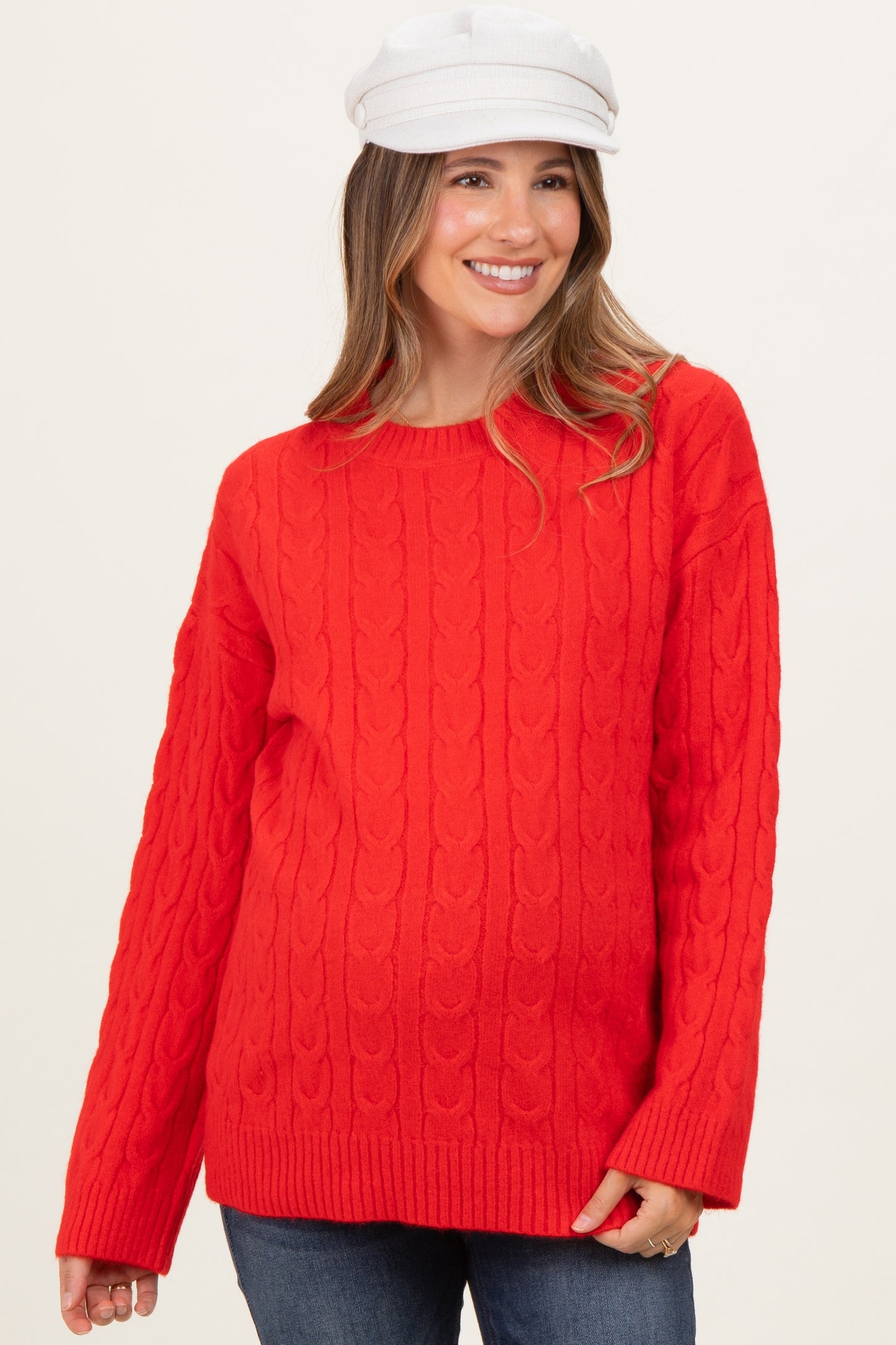 woman wearing a bright red cable knit maternity sweater with relaxed fit and long sleeves, smiling and wearing a white hat