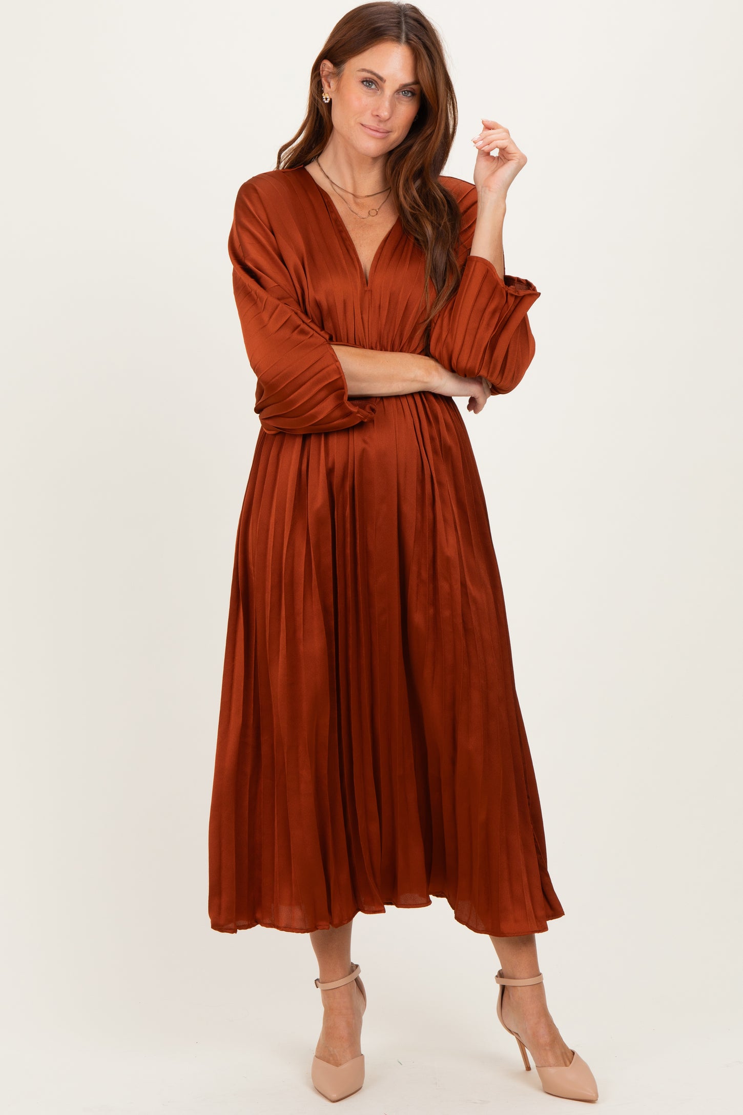 woman wearing an rust-colored pleated midi dress with long sleeves and a v-neckline, styled with beige heels