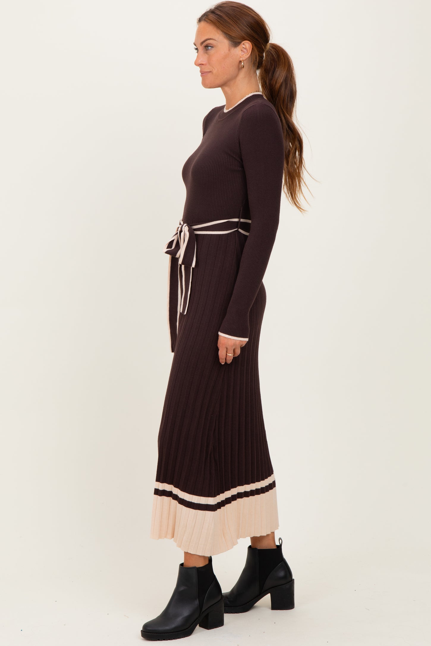 woman wearing a ribbed brown maxi dress with a beige pleated hem and a matching belt, styled with black ankle boots