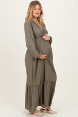 woman wearing a dark green maternity maxi dress with long sleeves and a gathered waist, holding her baby bump