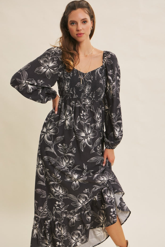 woman wearing a black floral maxi dress with long sleeves and a smocked bodice, posed against a neutral background