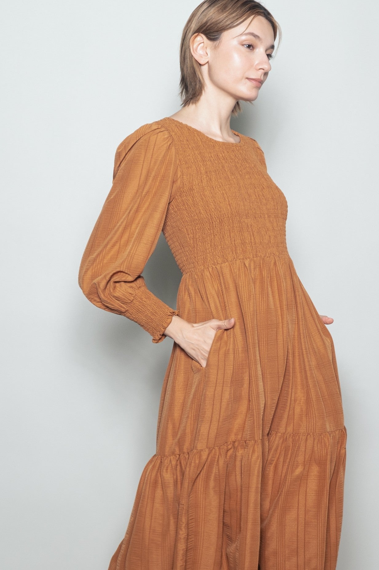 woman wearing a rust-colored long sleeve dress with a smocked bodice and tiered skirt, posing with hands in pockets