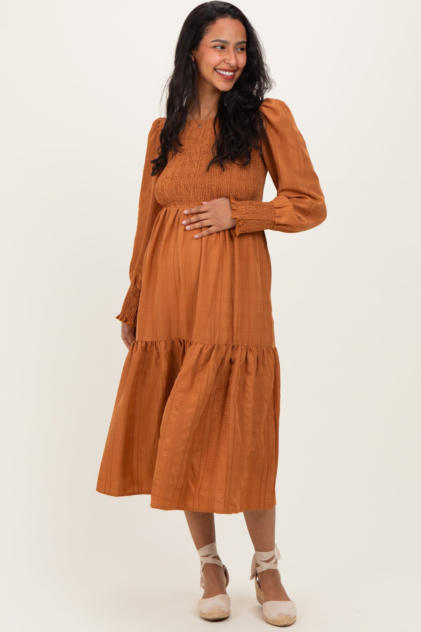 woman wearing a rust colored maternity midi dress with long sleeves and a smocked bodice, showing her baby bump