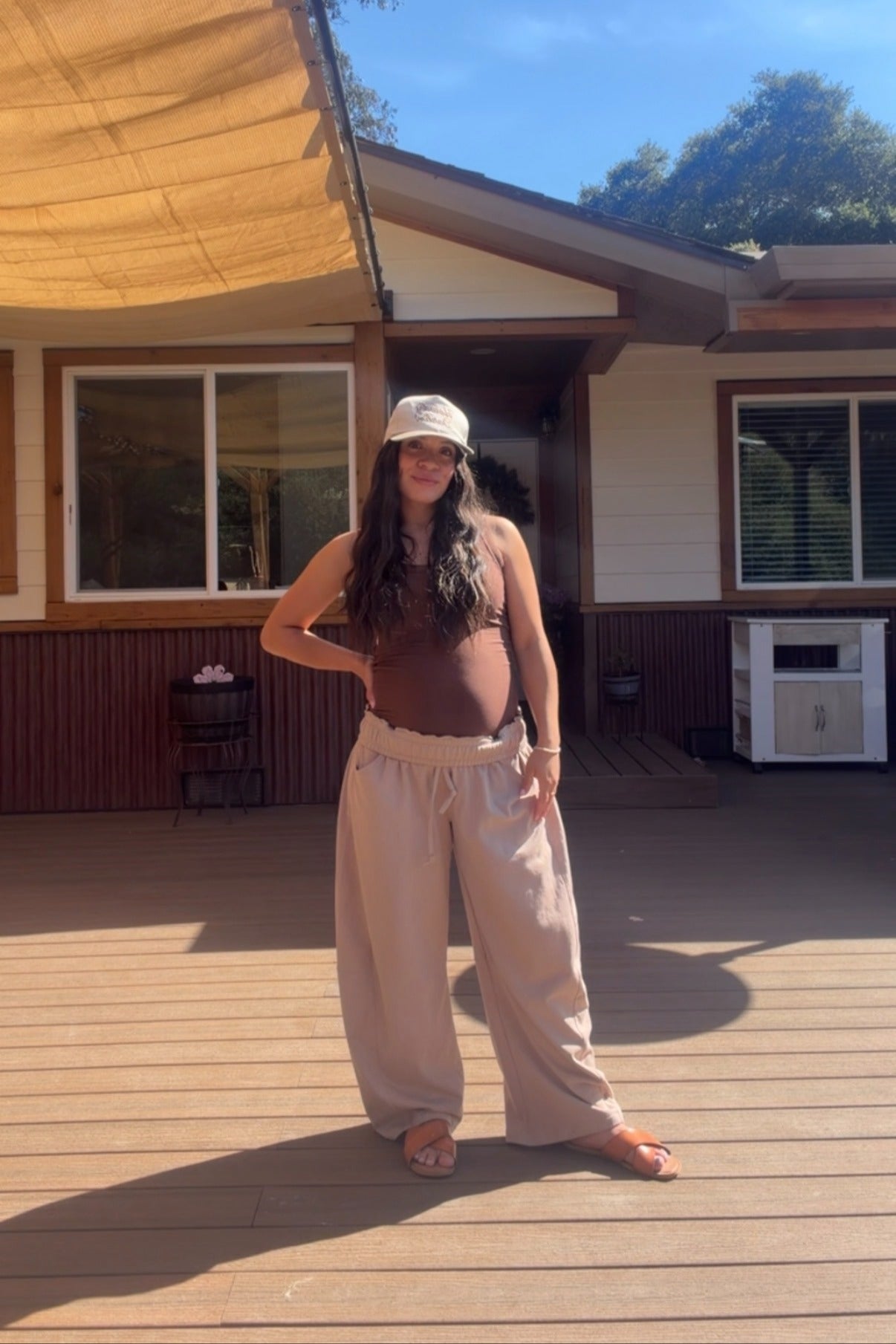 woman wearing a brown maternity tank top and loose beige maternity pants, showing baby bump, standing outdoors