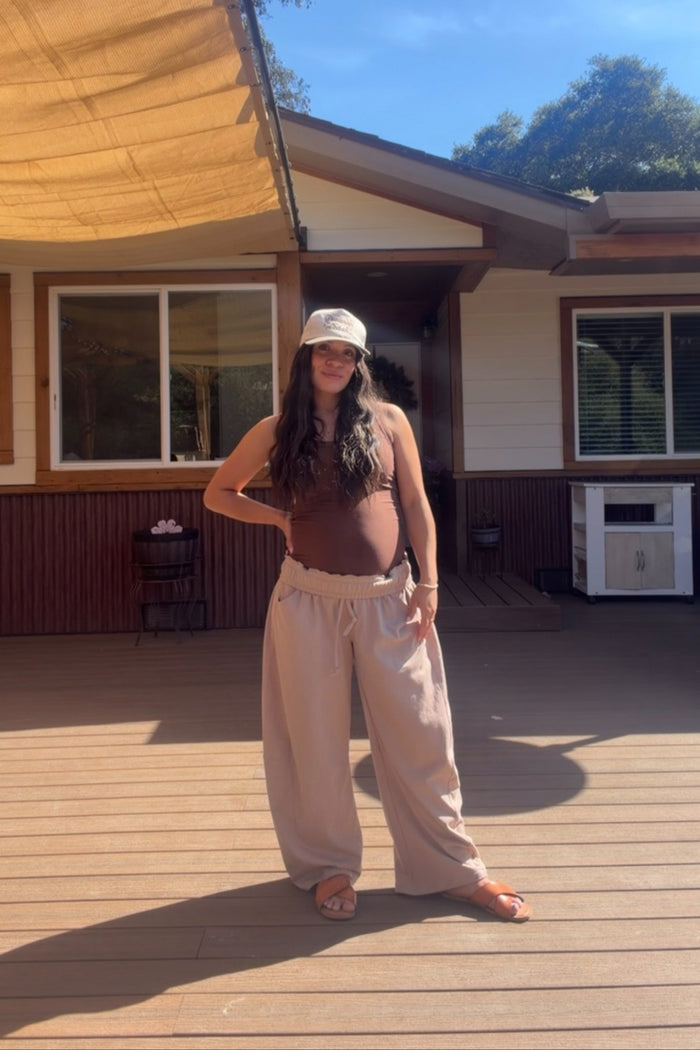 woman wearing a brown maternity tank top and loose beige maternity pants, showing baby bump, standing outdoors