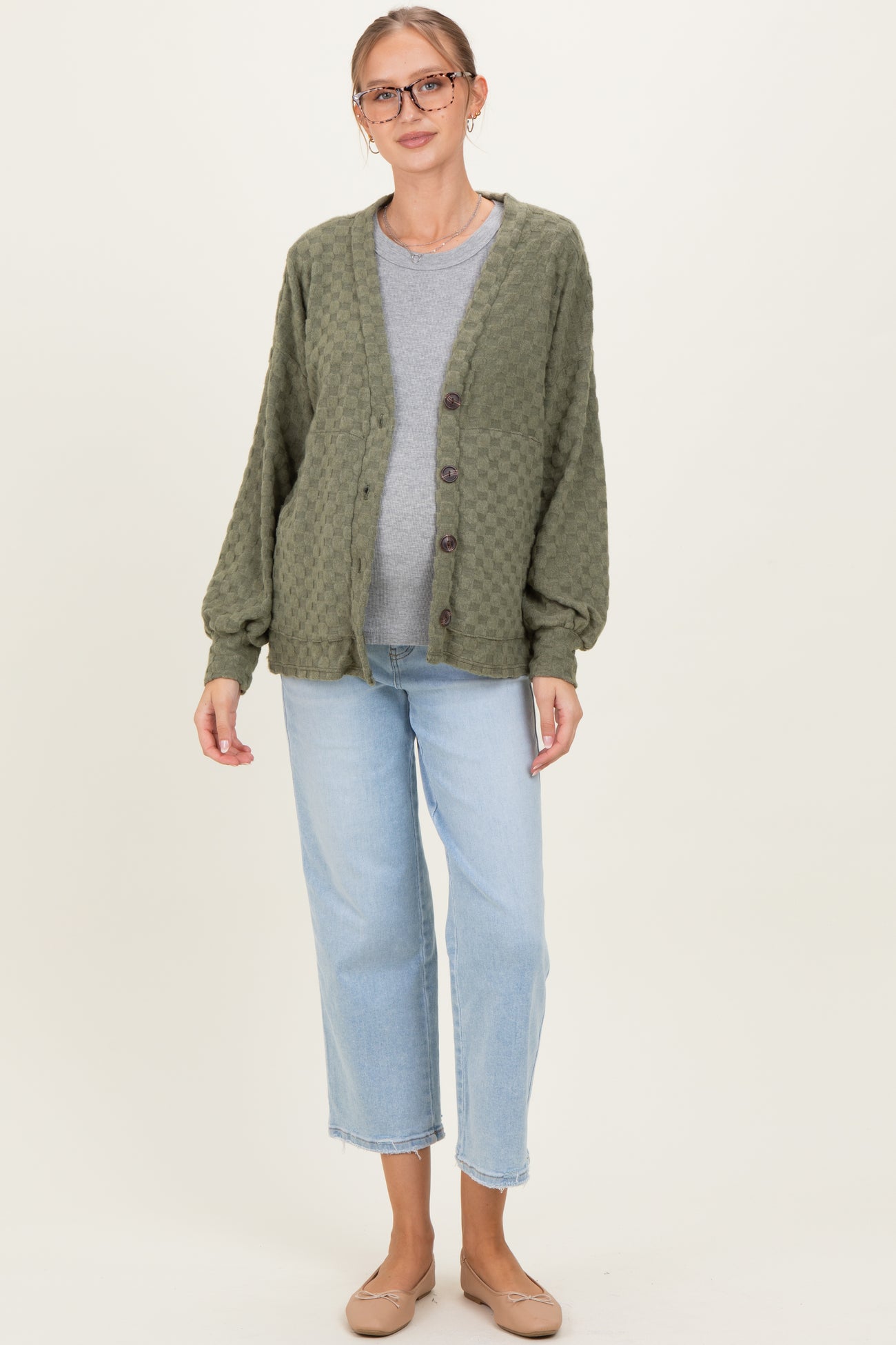woman wearing a green textured maternity cardigan over a gray shirt and light blue jeans, smiling with a baby bump visible