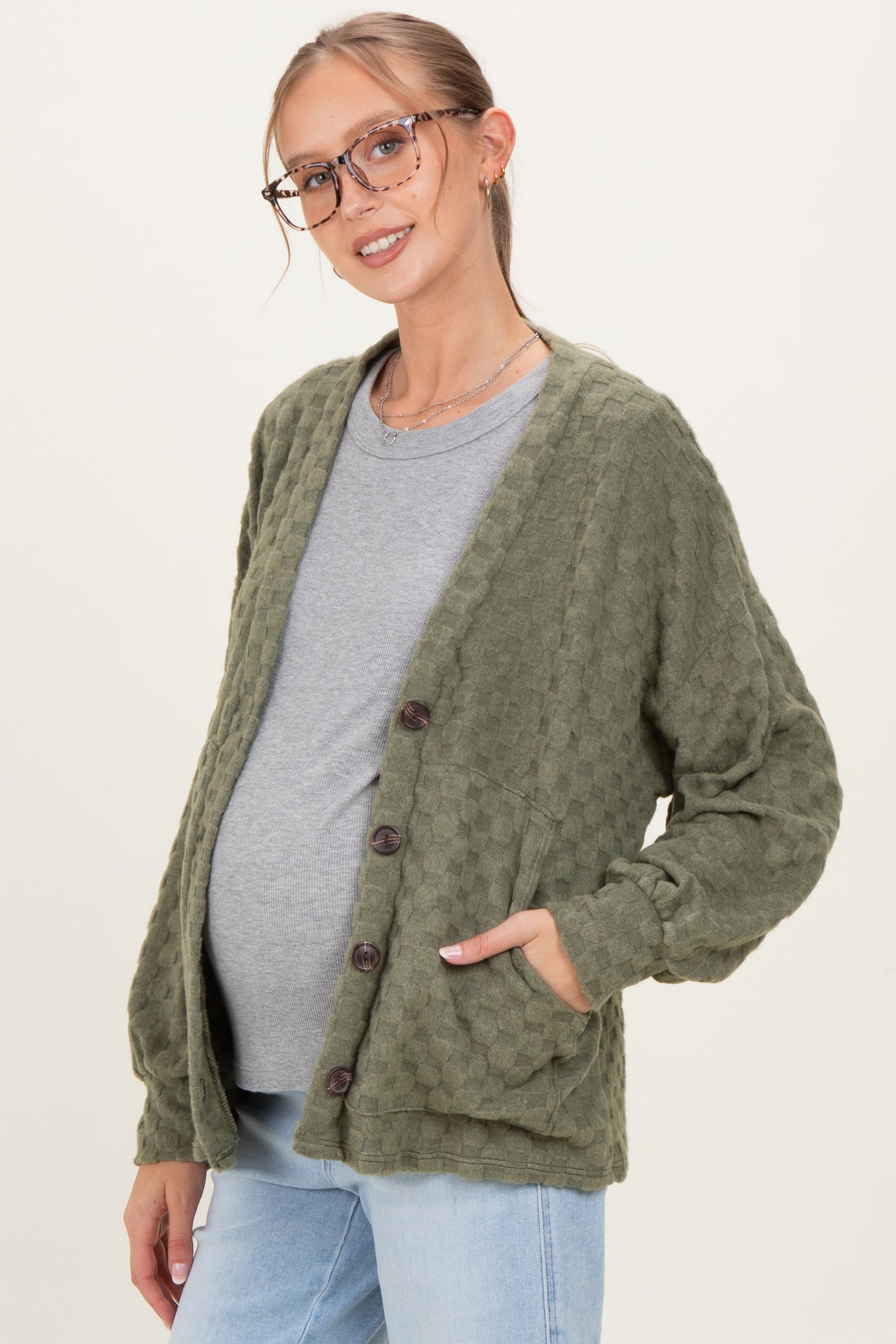 woman wearing an olive green textured maternity cardigan over a gray top, hands in pockets, showcasing a baby bump