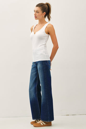 woman wearing a white v-neck camisole top and wide leg blue denim jeans, standing with hands on hips