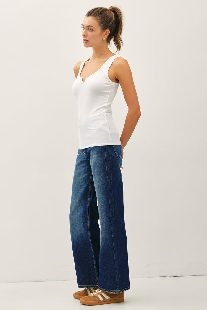 woman wearing a white v-neck camisole top and wide leg blue denim jeans, standing with hands on hips
