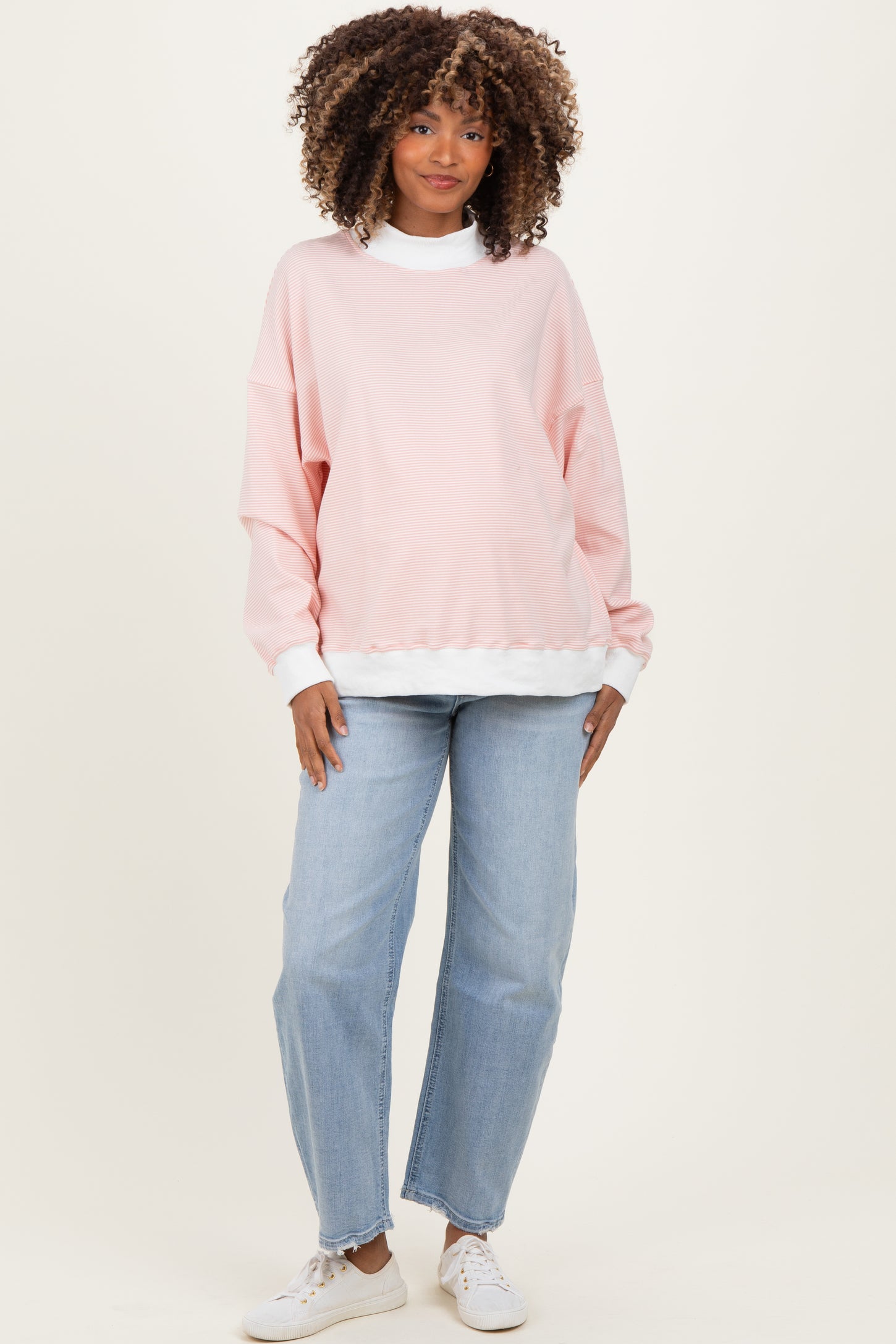 woman wearing a light pink oversized maternity sweatshirt with a white trim and blue jeans, smiling at the camera