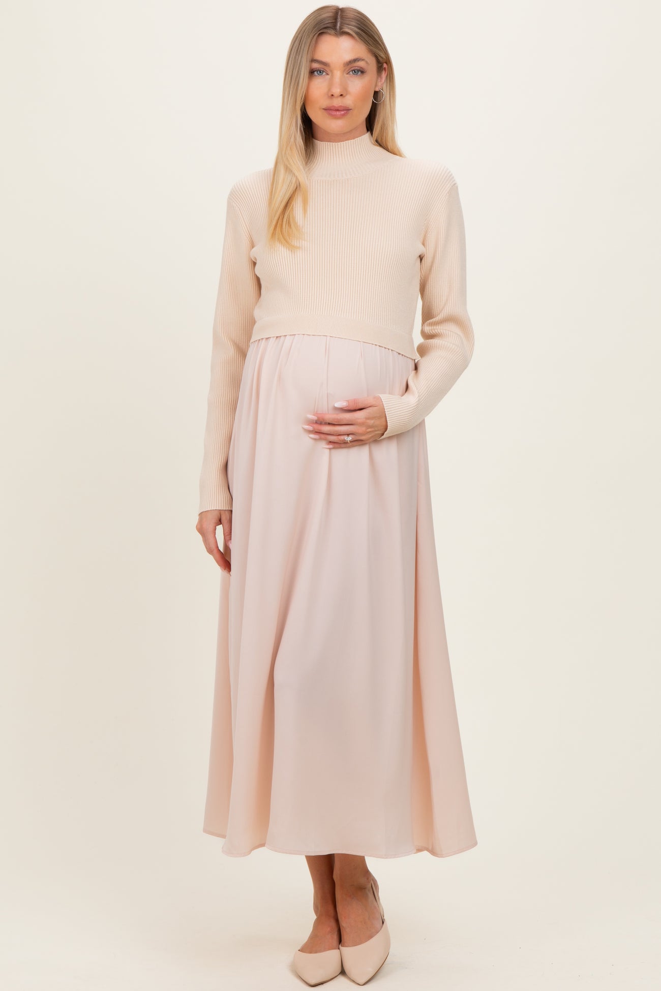 woman wearing a cream ribbed maternity sweater and a pale pink maxi skirt, gently holding her baby bump