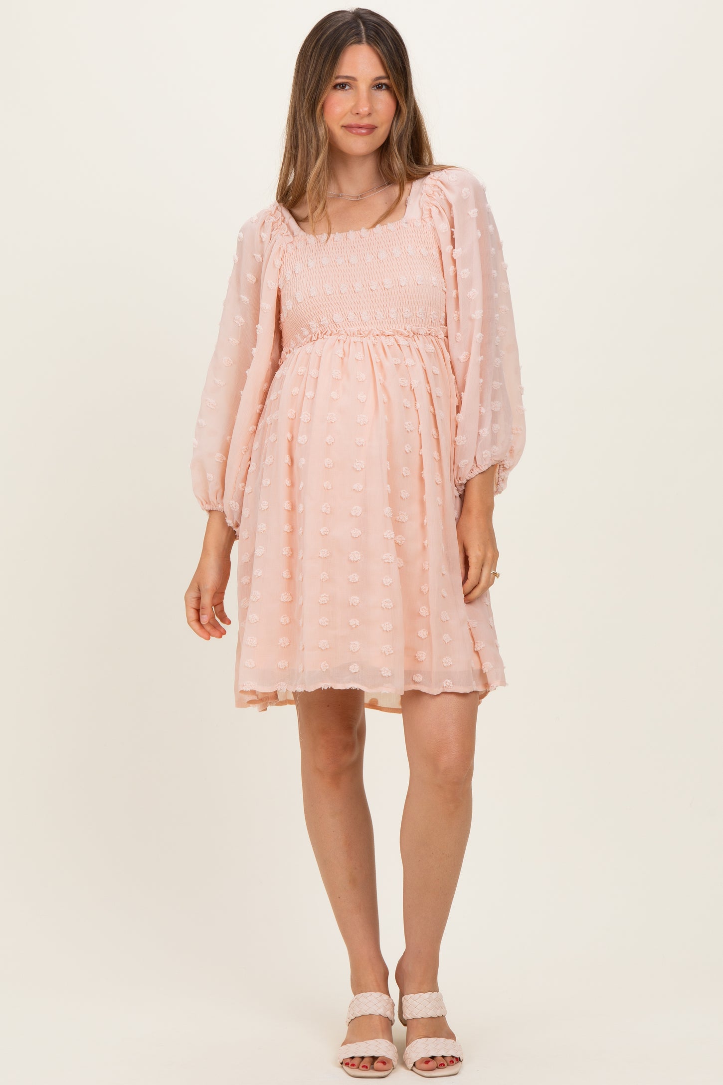woman wearing a light pink maternity dress with puff sleeves and a smocked bodice, standing in a relaxed pose
