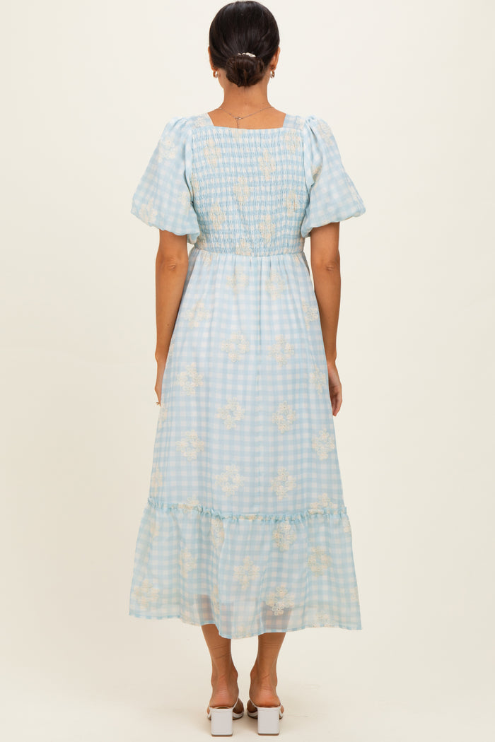 woman wearing a light blue gingham patterned maxi dress with short puff sleeves and floral detailing