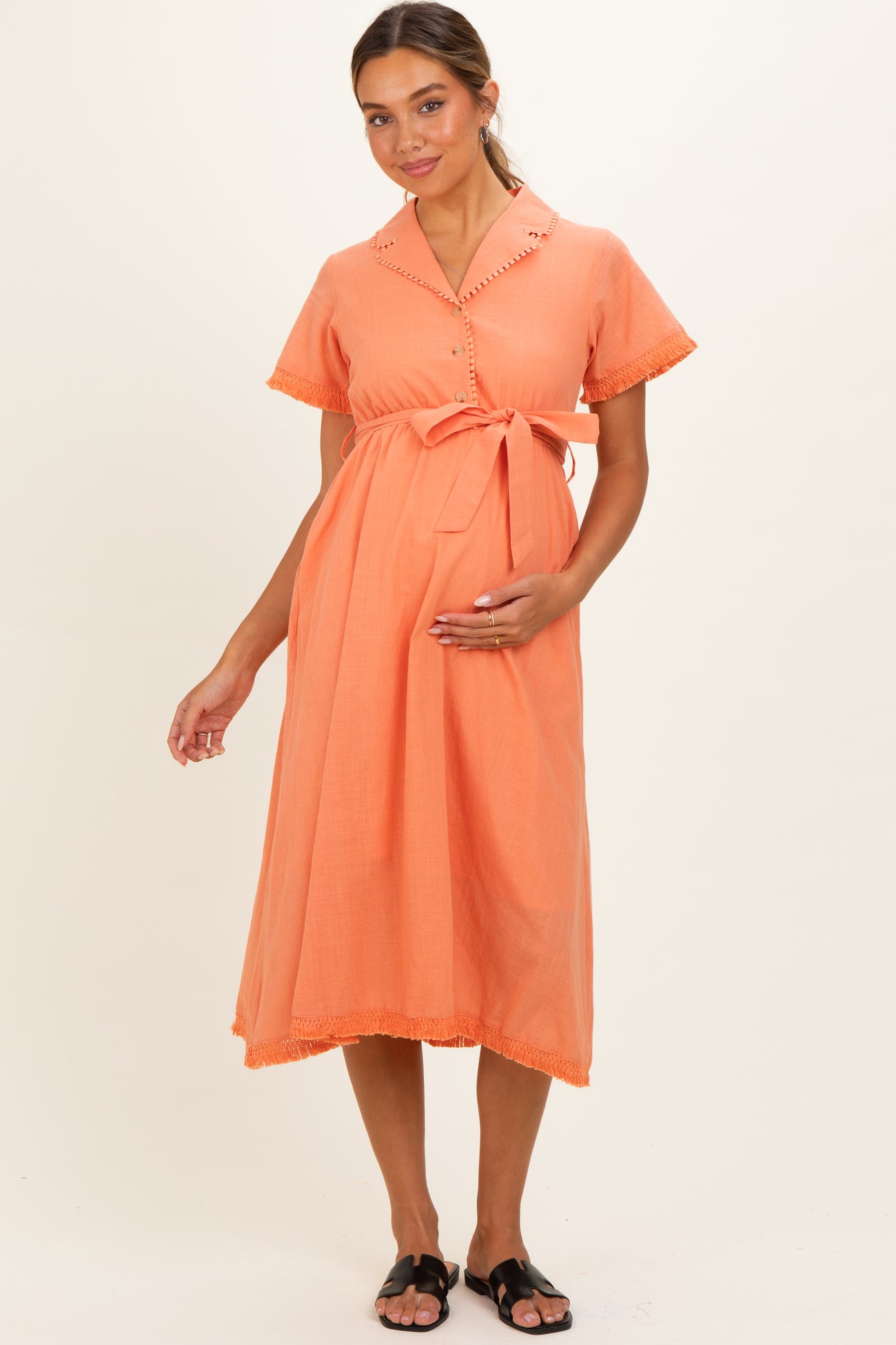 woman wearing a coral maternity dress with short sleeves, button details, and a tie belt, holding her baby bump