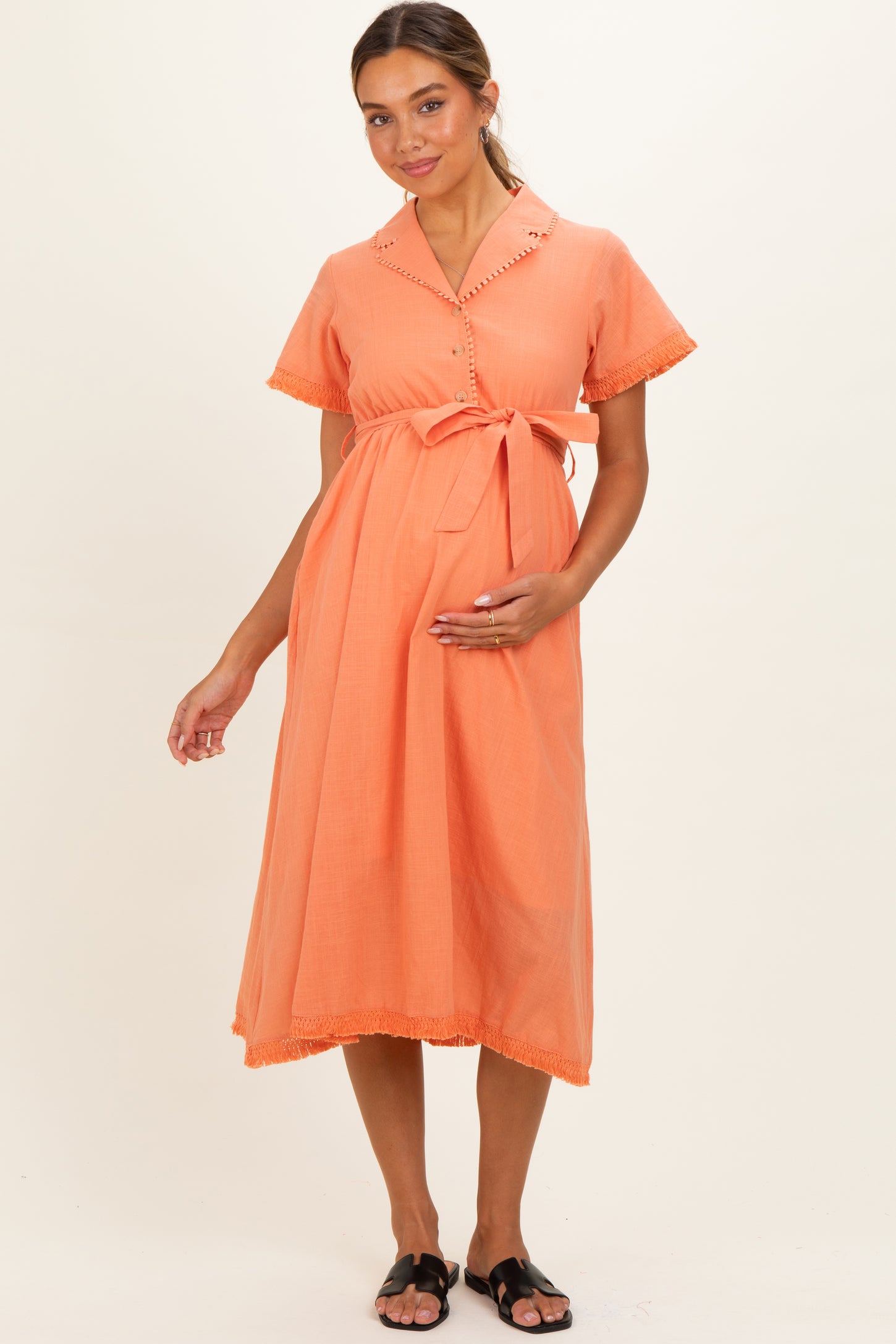 woman wearing a coral maternity dress with short sleeves, button details, and a tie belt, holding her baby bump
