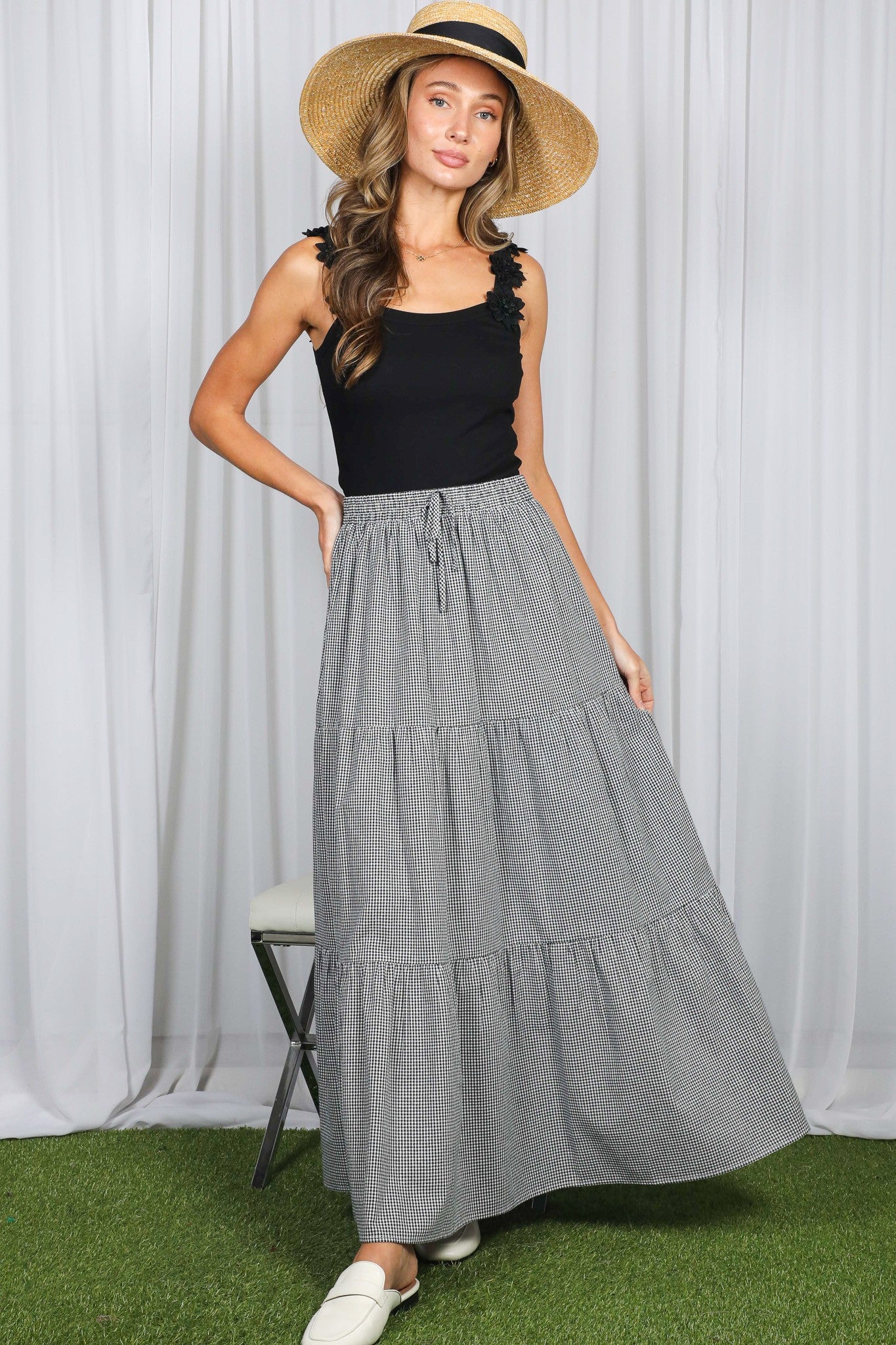 woman wearing a black tank top and a black and white striped tiered maxi skirt, standing with a straw hat