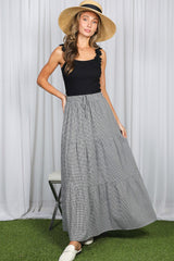 woman wearing a black tank top and a black and white striped tiered maxi skirt, standing with a straw hat