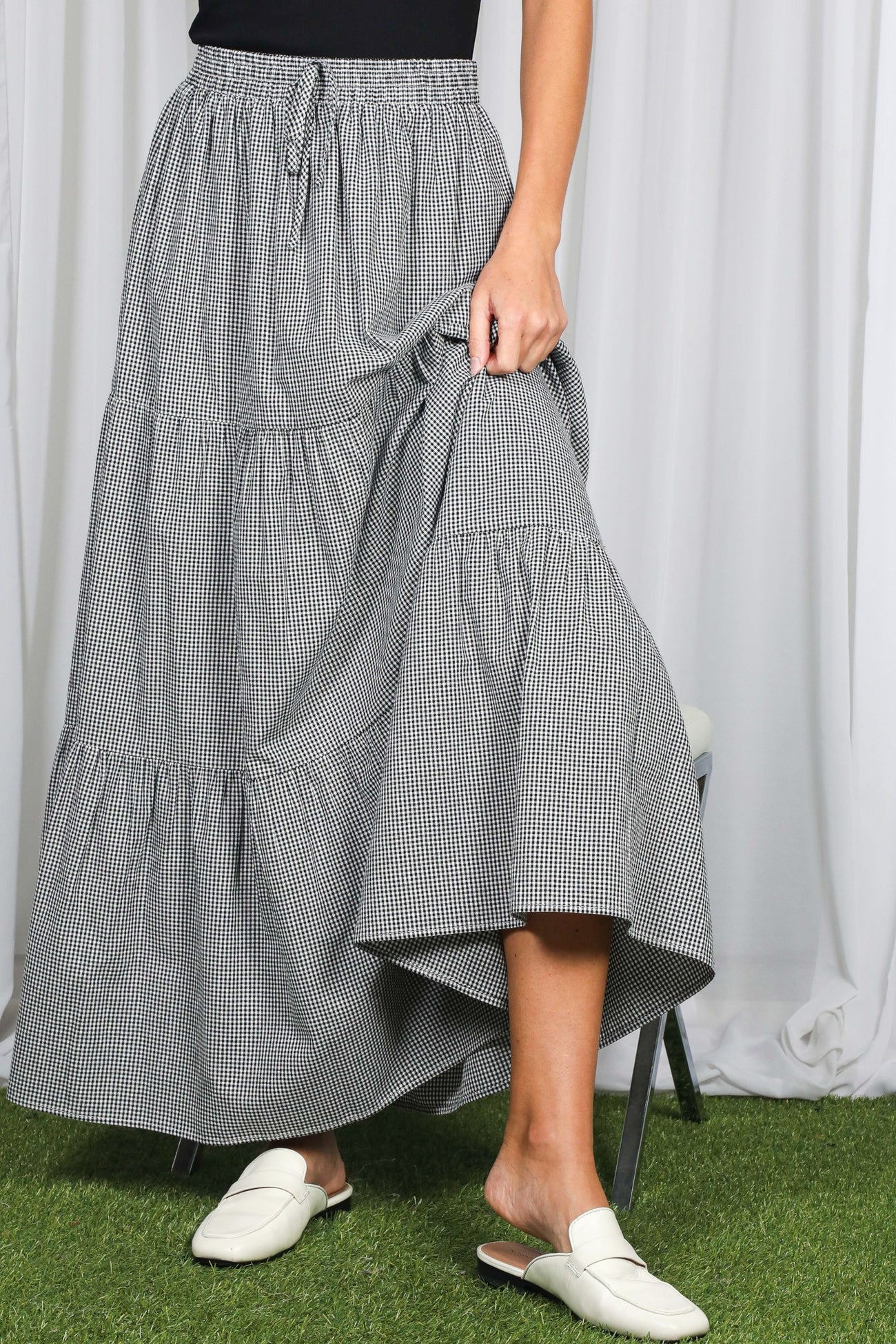 woman wearing a black and white gingham tiered maxi skirt and white slide sandals, holding the skirt hem