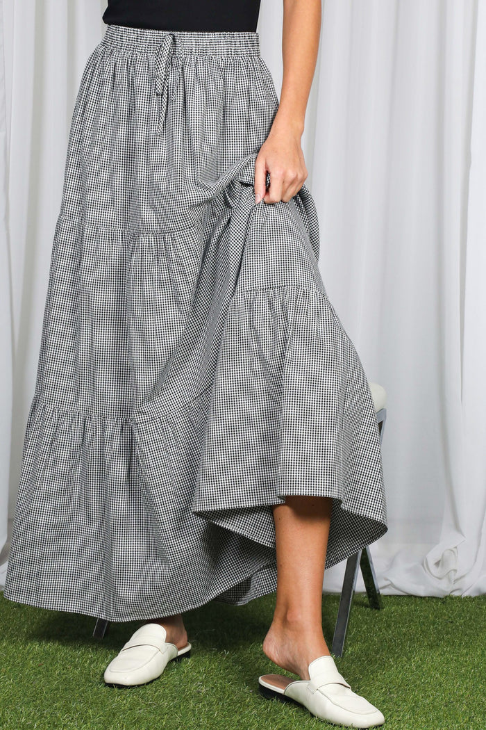 woman wearing a black and white gingham tiered maxi skirt and white slide sandals, holding the skirt hem