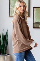 woman wearing a brown oversized ribbed sweater with cream cuff details and blue jeans, standing in a bright room