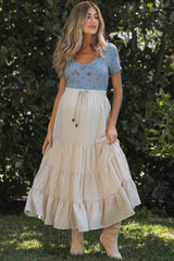 woman wearing a blue embroidered maternity top and a cream tiered maxi skirt, standing in a garden setting