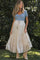 woman wearing a blue embroidered maternity top and a cream tiered maxi skirt, standing in a garden setting
