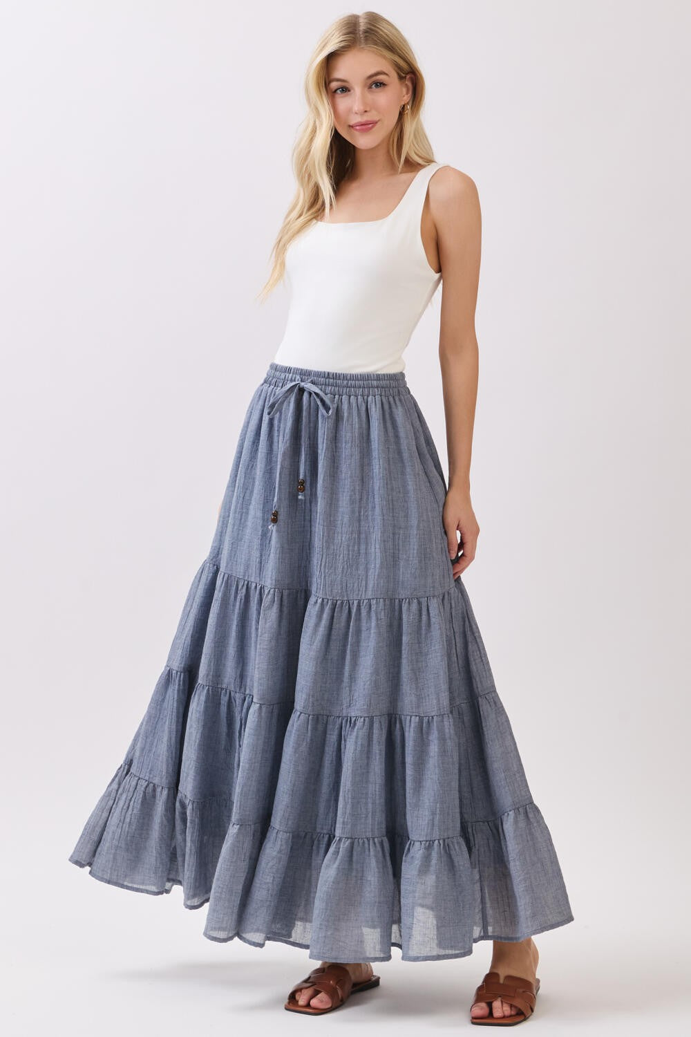 woman wearing a white tank top and a tiered blue maxi skirt, standing with one hand on her hip