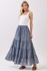 woman wearing a white tank top and a tiered blue maxi skirt, standing with one hand on her hip