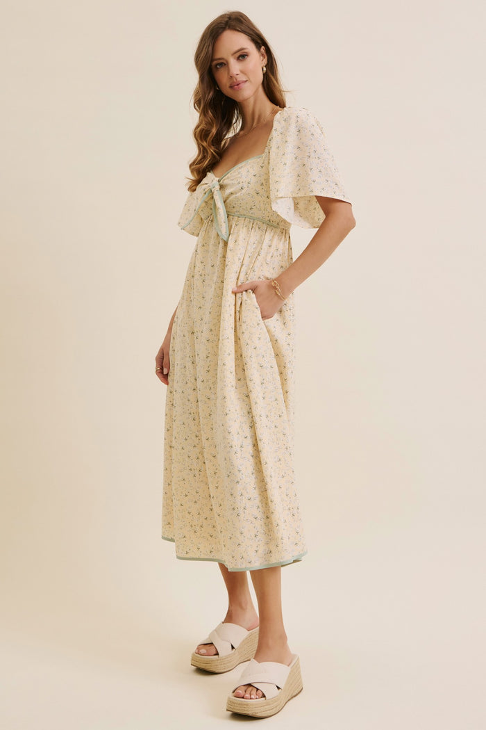 woman wearing a floral print yellow maxi dress with a tie-front detail and flutter sleeves, paired with white wedge sandals