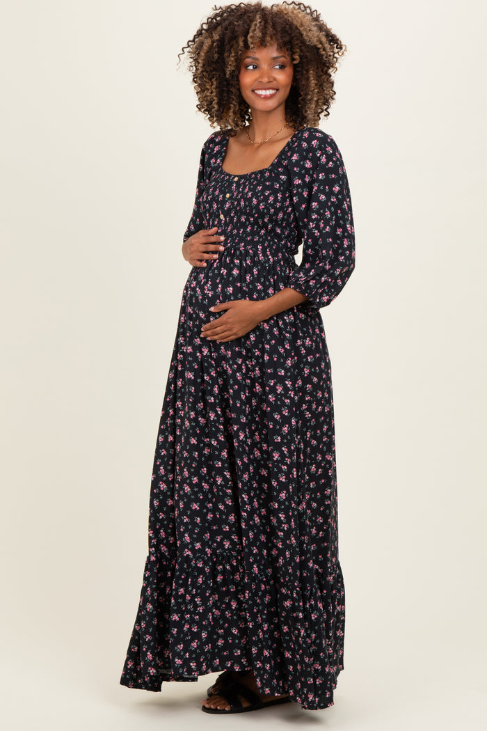 woman wearing a black floral maternity maxi dress with a square neckline and three-quarter sleeves, holding her baby bump